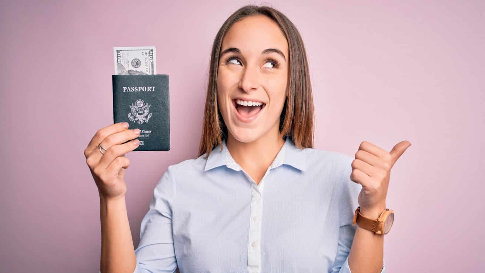Smiling woman holding a passport with cash inside and giving a thumbs up against a pink background.
