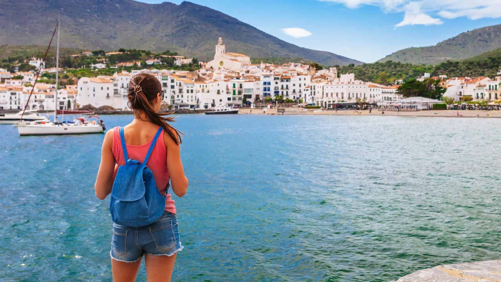 Woman with a backpack stands by the water, looking at a coastal town with mountains in the background.