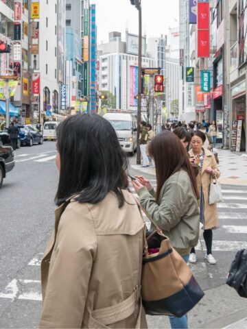 People waiting at a busy crosswalk in a city with shops and cars in the background.