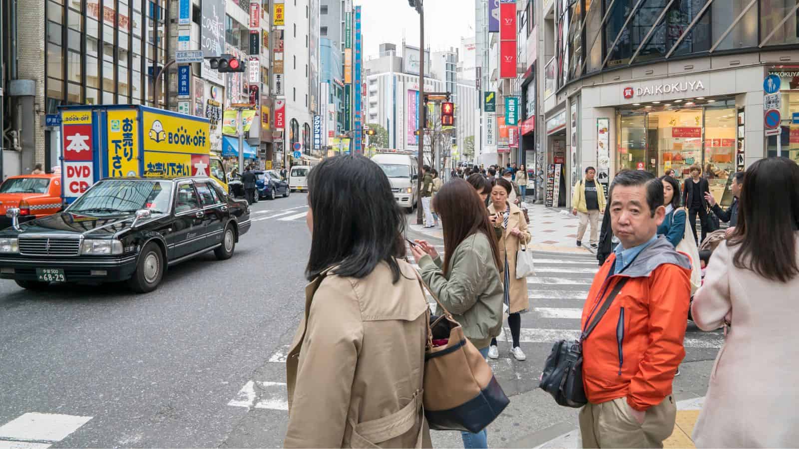 People waiting at a busy crosswalk in a city with shops and cars in the background.
