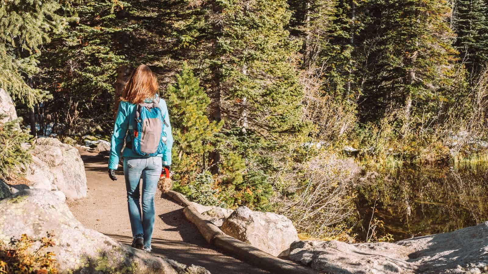 A woman with a backpack hiking on a forest trail surrounded by rocks and tall trees.