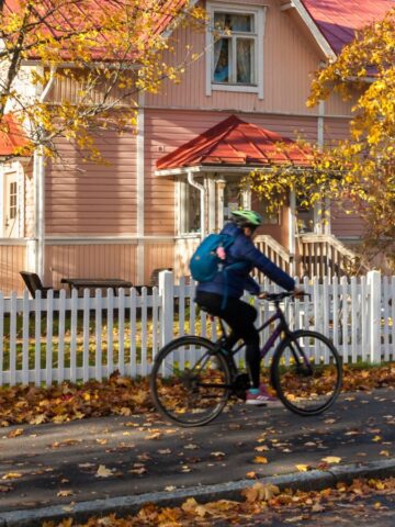 A person rides a bicycle past a pink house with a white fence in a small town, surrounded by autumn trees and fallen leaves.