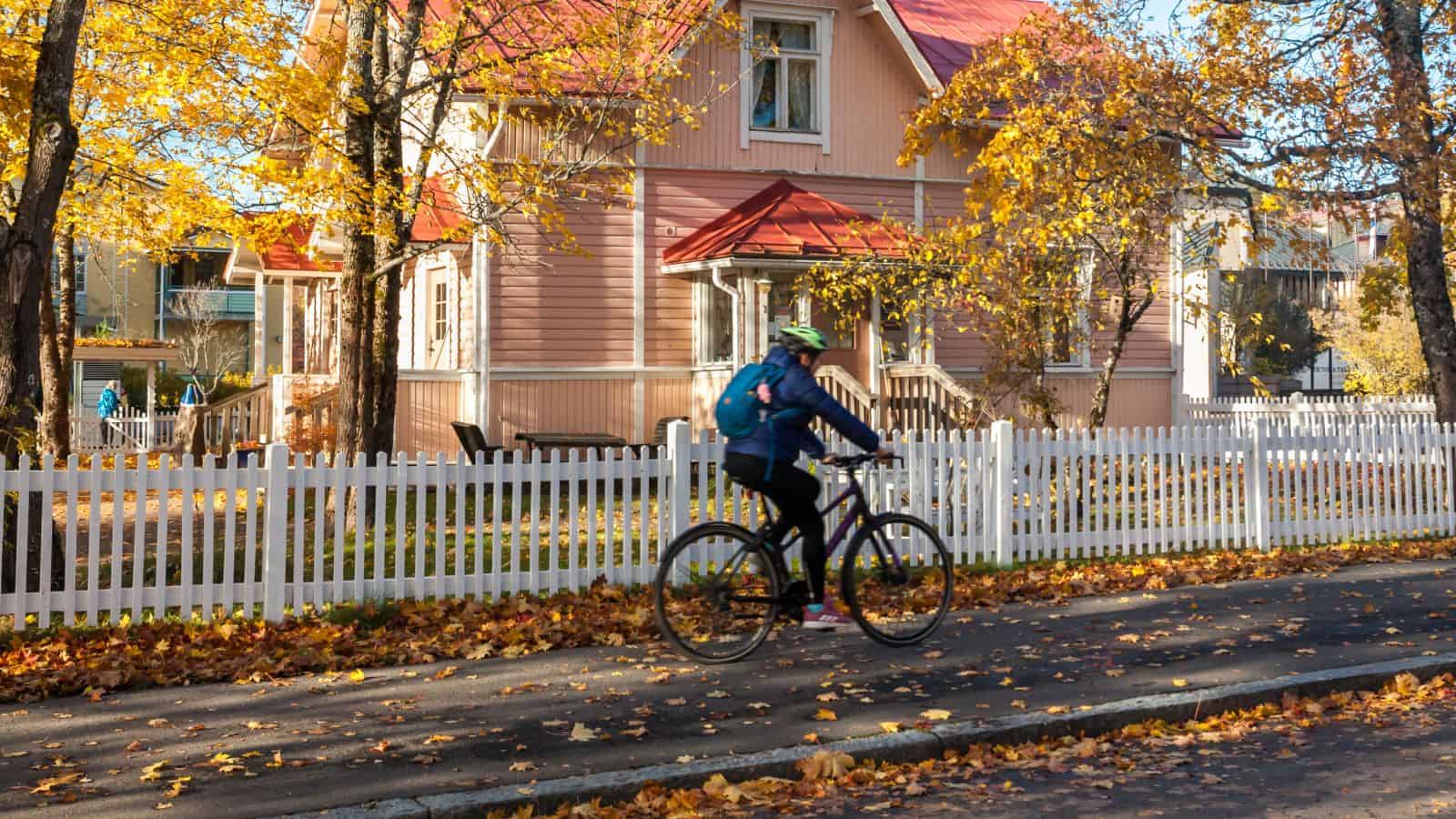 A person rides a bicycle past a pink house with a white fence in a small town, surrounded by autumn trees and fallen leaves.