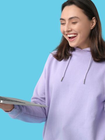 Smiling woman in a lavender hoodie holding a laptop and gesturing, standing against a blue background.