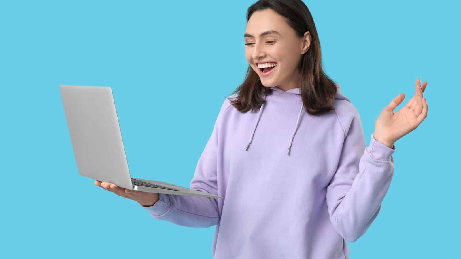 Smiling woman in a lavender hoodie holding a laptop and gesturing, standing against a blue background.