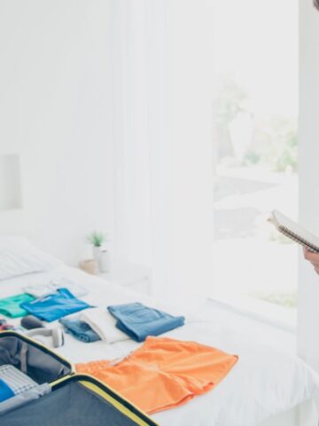 Man thinking and holding a notepad while packing clothes and accessories into a suitcase on a bed.