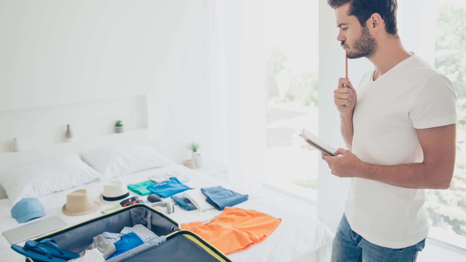 Man thinking and holding a notepad while packing clothes and accessories into a suitcase on a bed.