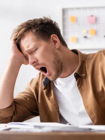 A tired man yawning at a desk with a laptop, papers, and a coffee cup in front of him.