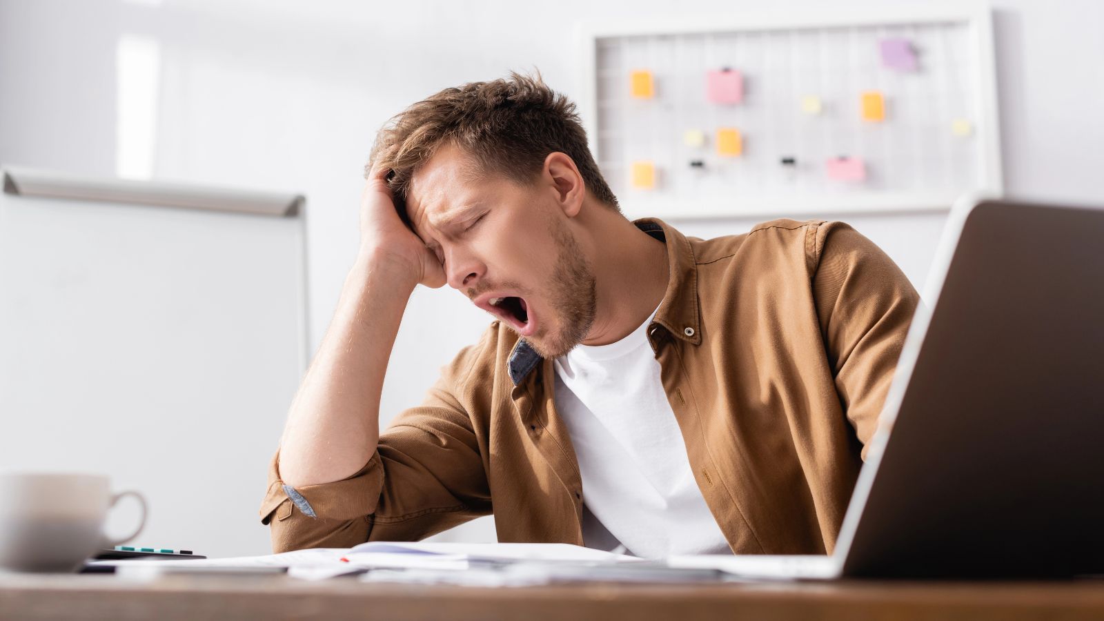 A tired man yawning at a desk with a laptop, papers, and a coffee cup in front of him.