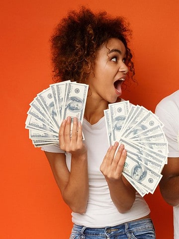 Two excited people holding and fanning out lots of cash against an orange background, ready to share their favorite ways to save money while you’re already on a cruise.