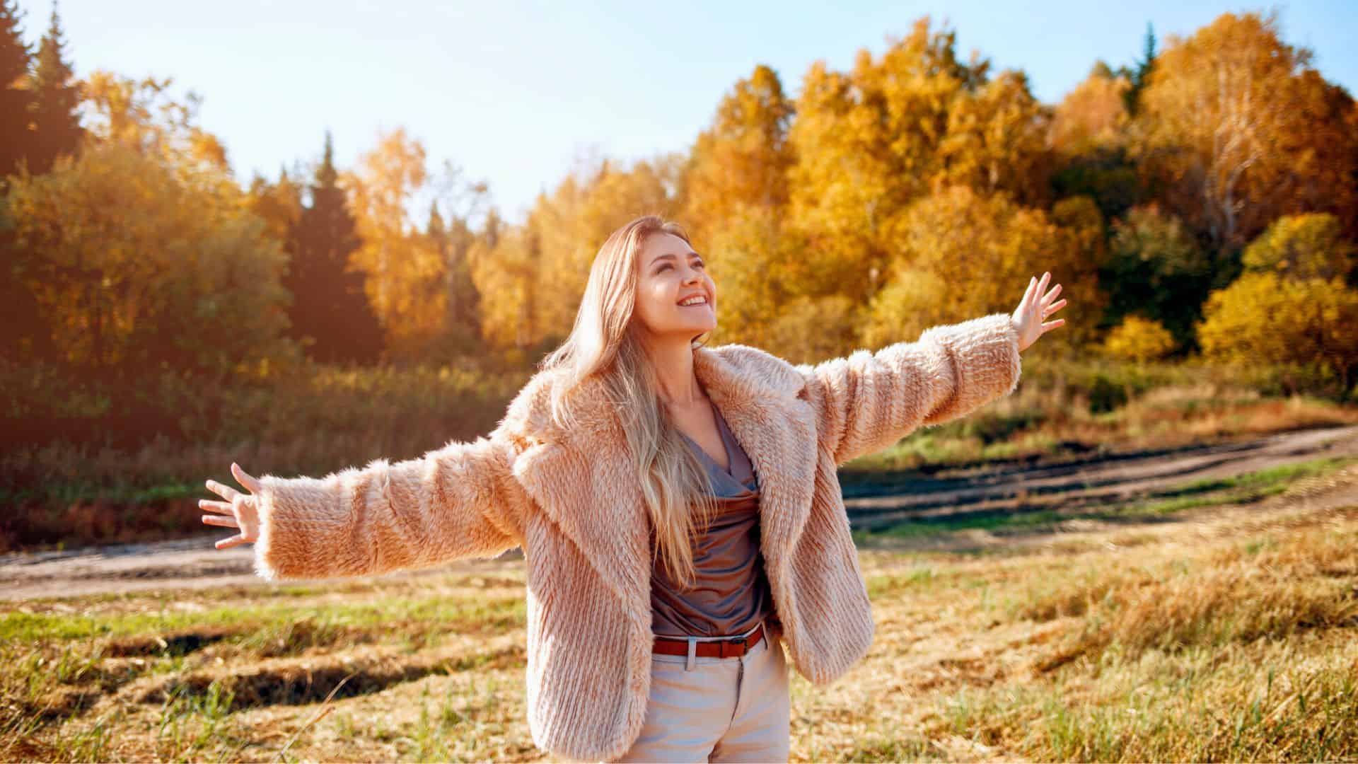 Smiling woman in a fur coat stands outdoors with arms outstretched, surrounded by autumn trees.