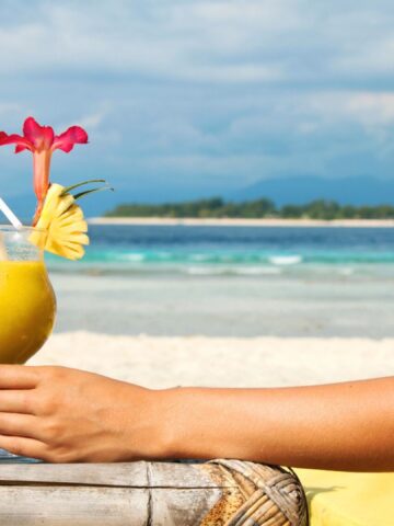 A person relaxing on a beach chair holding a tropical drink with a flower and pineapple garnish by the ocean.