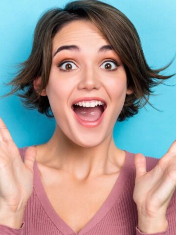 Woman with short brown hair smiling widely and holding hands up in excitement against a blue background.