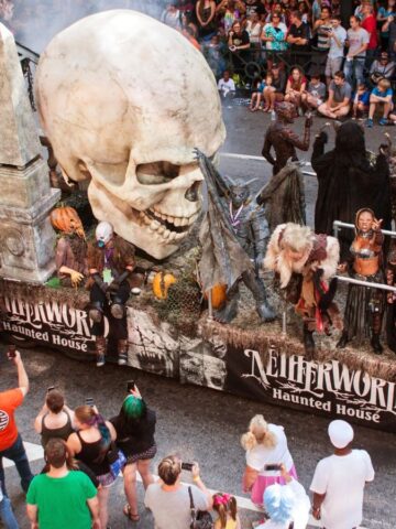 Parade float with a giant skull, spooky figures, and "Netherworld Haunted House" sign, surrounded by crowds.