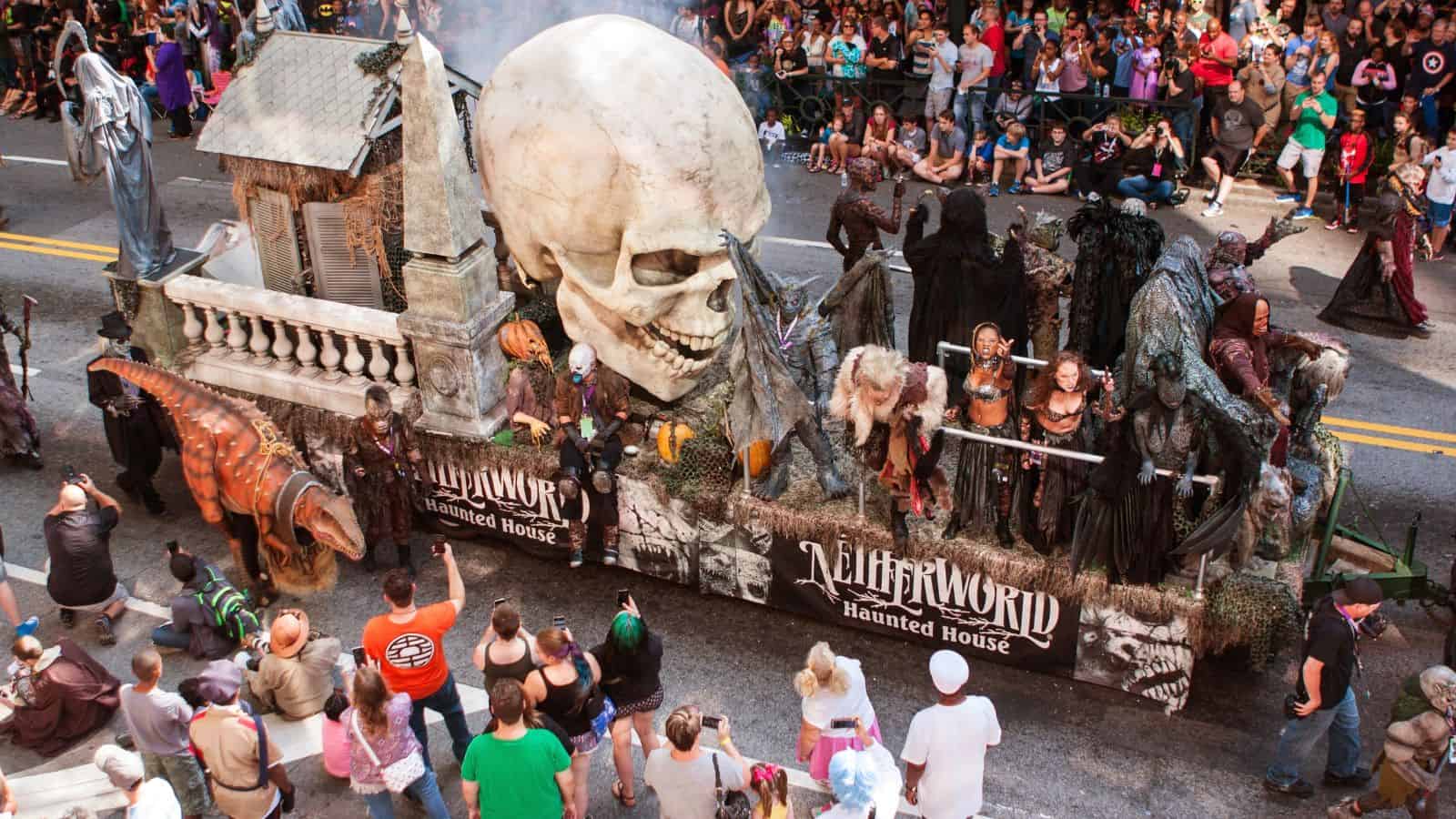 Parade float with a giant skull, spooky figures, and "Netherworld Haunted House" sign, surrounded by crowds.