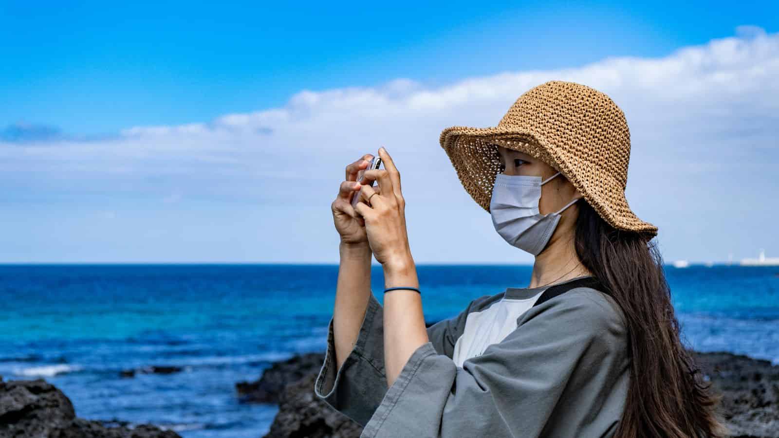 Woman in a straw hat and mask taking a photo by the ocean under a blue sky.