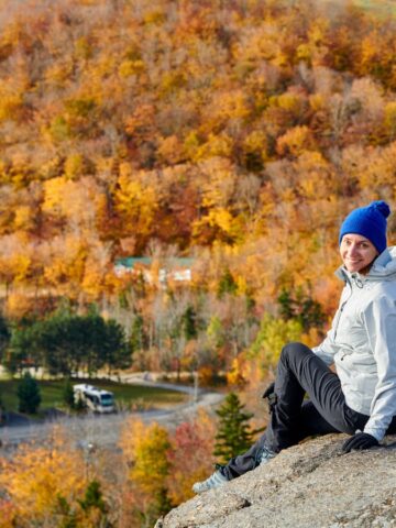 Person in a blue hat sits on a rocky ledge with colorful autumn trees in the background, enjoying one of the perfect hidden destinations for weekend getaways.