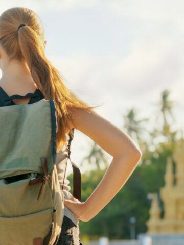 A woman with a backpack stands facing a golden temple under a bright sky.