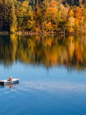 A person rows a small boat on a calm lake with colorful autumn trees reflected in the water.