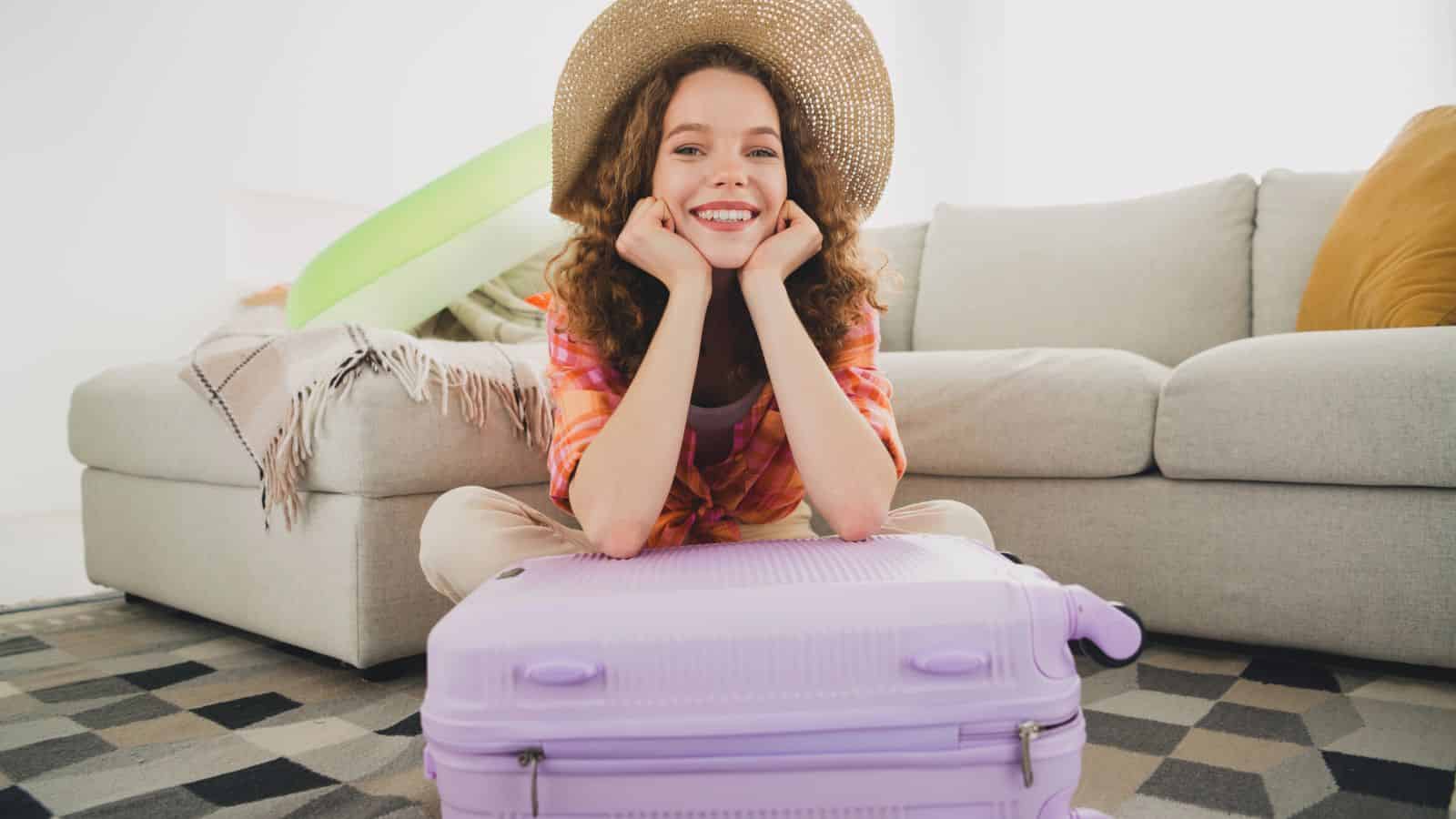 Smiling woman in a hat sits behind a purple suitcase in a living room, ready for travel.