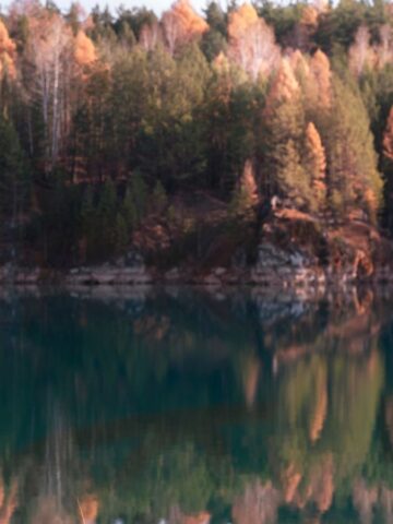 Person in a jacket and beanie uses binoculars by a lake, with autumn trees reflected in the water.