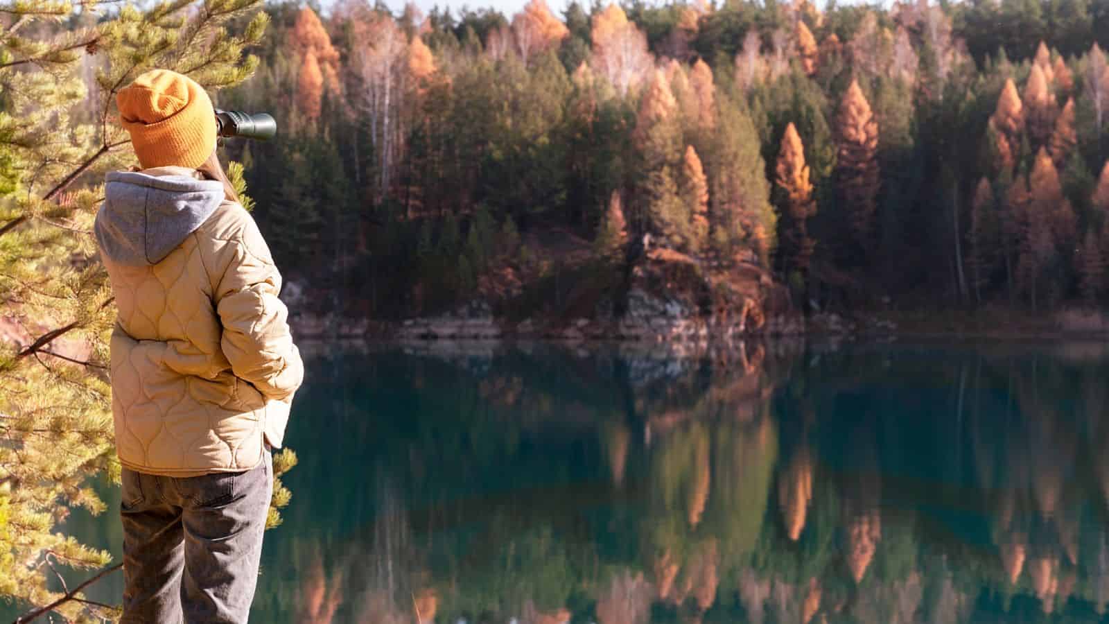 Person in a jacket and beanie uses binoculars by a lake, with autumn trees reflected in the water.