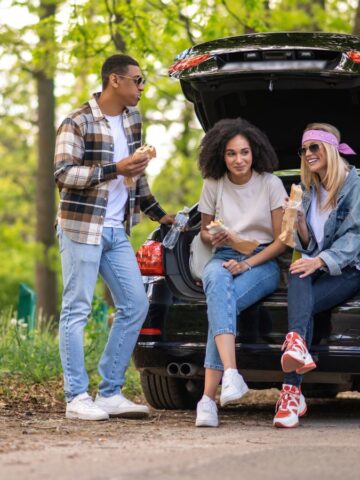 Four friends share snacks and laugh while sitting by an open car trunk on a forested roadside.
