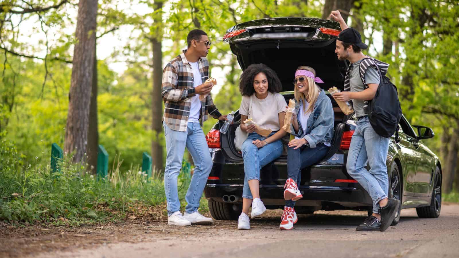 Four friends share snacks and laugh while sitting by an open car trunk on a forested roadside.