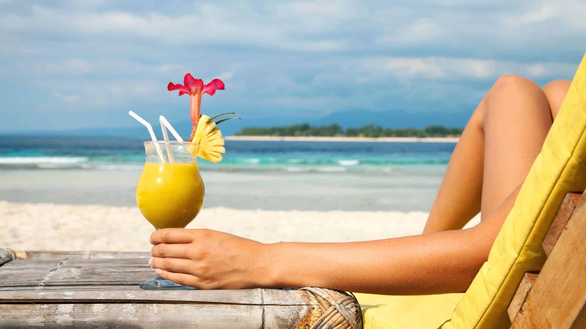 A person relaxing on a beach chair holding a tropical drink with a flower and pineapple garnish by the ocean.