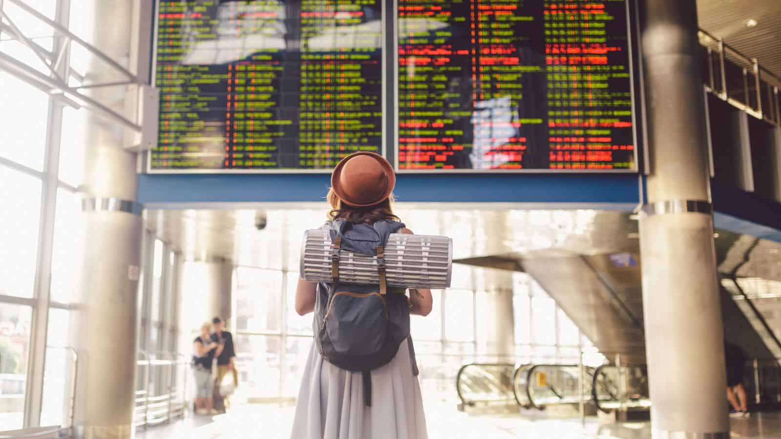 A traveler with a backpack and hat looks at a large flight information board in an airport terminal.