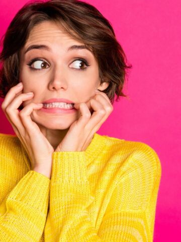 A girl in a yellow sweater looking scared biting her pinkies.