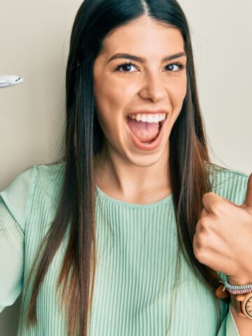 Smiling woman holding a toy airplane and giving a thumbs up against a plain background.