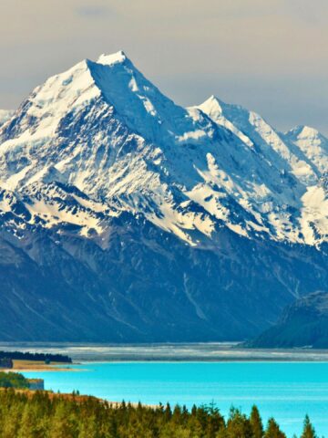 Snow-capped mountains rise above a turquoise lake and green forest under a partly cloudy sky.