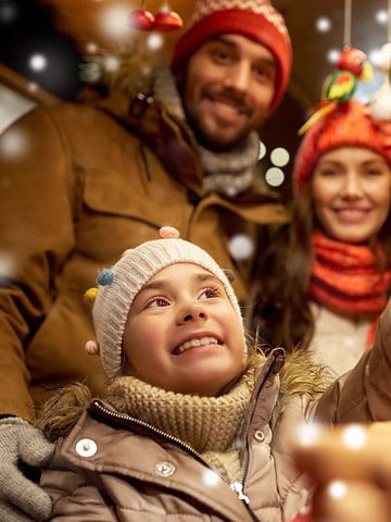 Smiling family in winter clothes shopping at a festive market with snow falling and decorations around.