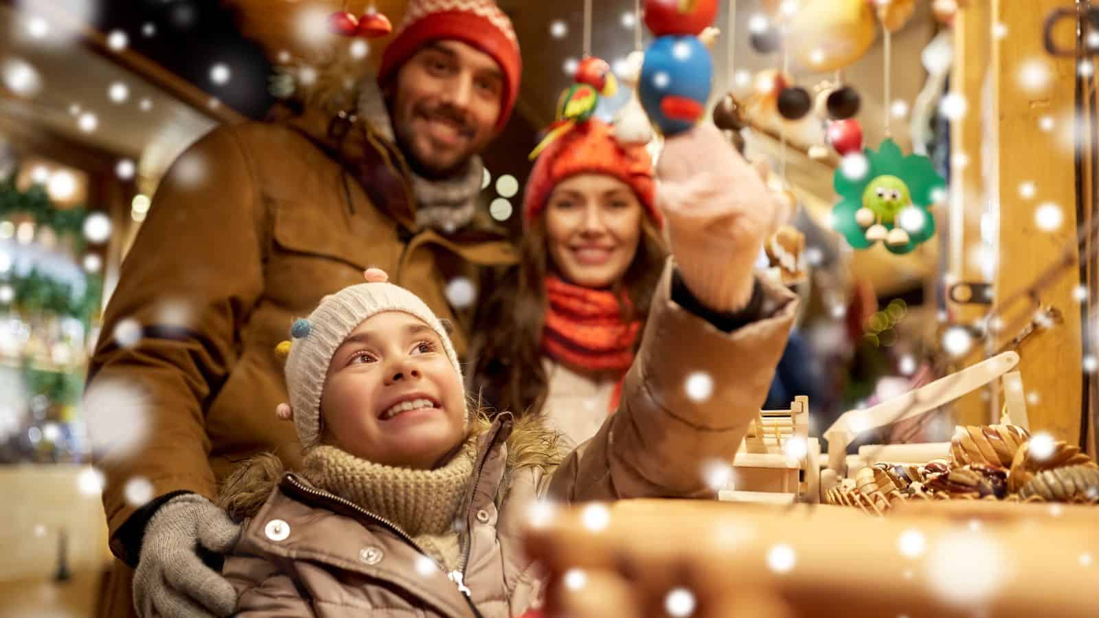 Smiling family in winter clothes shopping at a festive market with snow falling and decorations around.