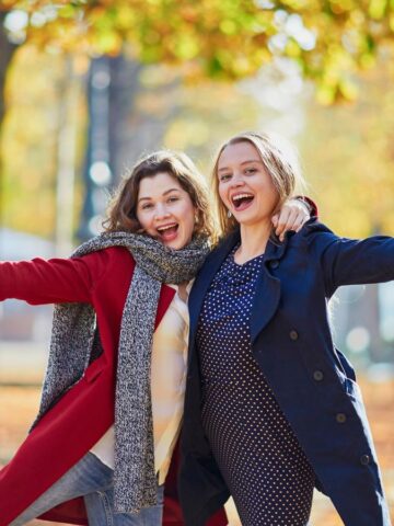 Two women in autumn coats smile and pose with outstretched arms in a park with fall foliage.