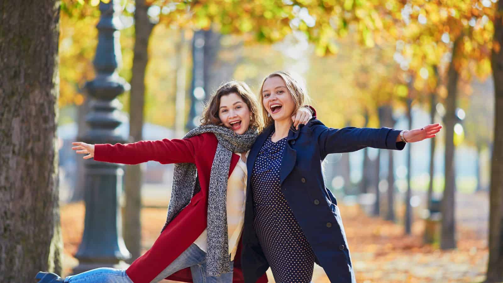 Two women in autumn coats smile and pose with outstretched arms in a park with fall foliage.