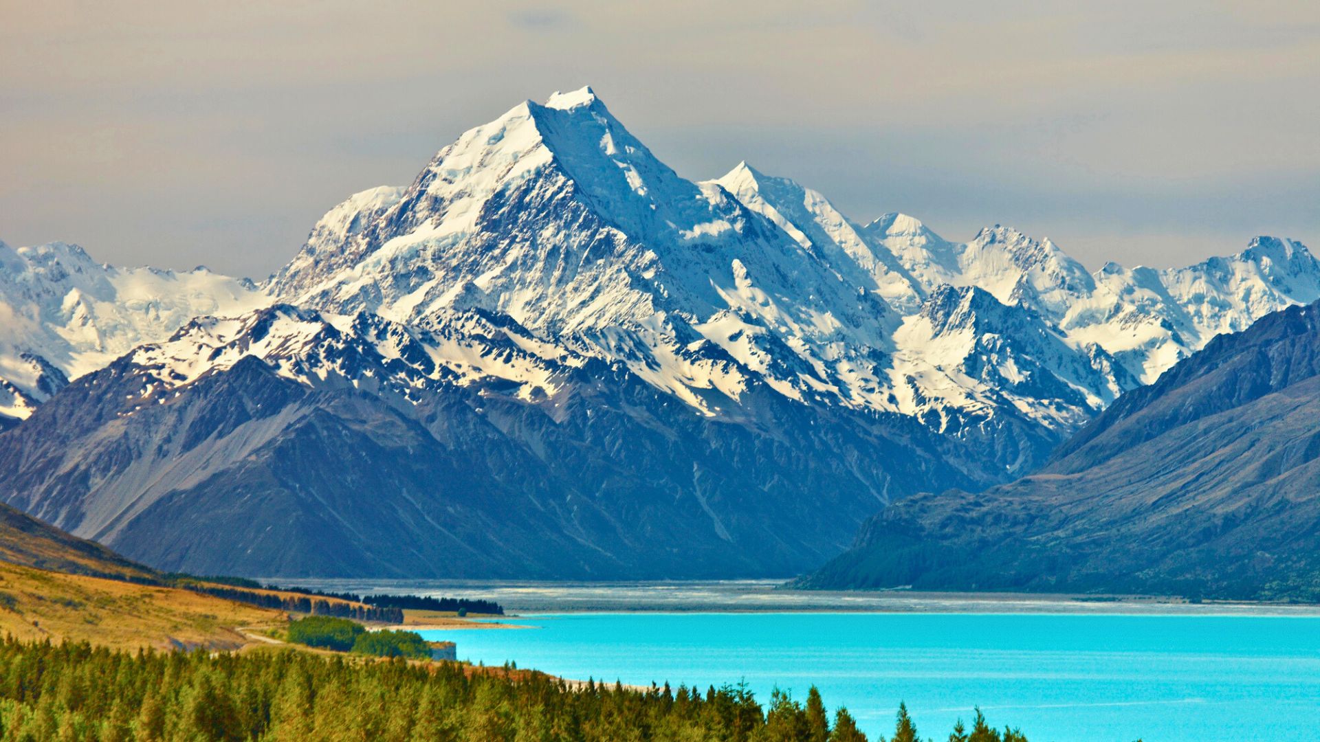 Snow-capped mountains rise above a turquoise lake and green forest under a partly cloudy sky.