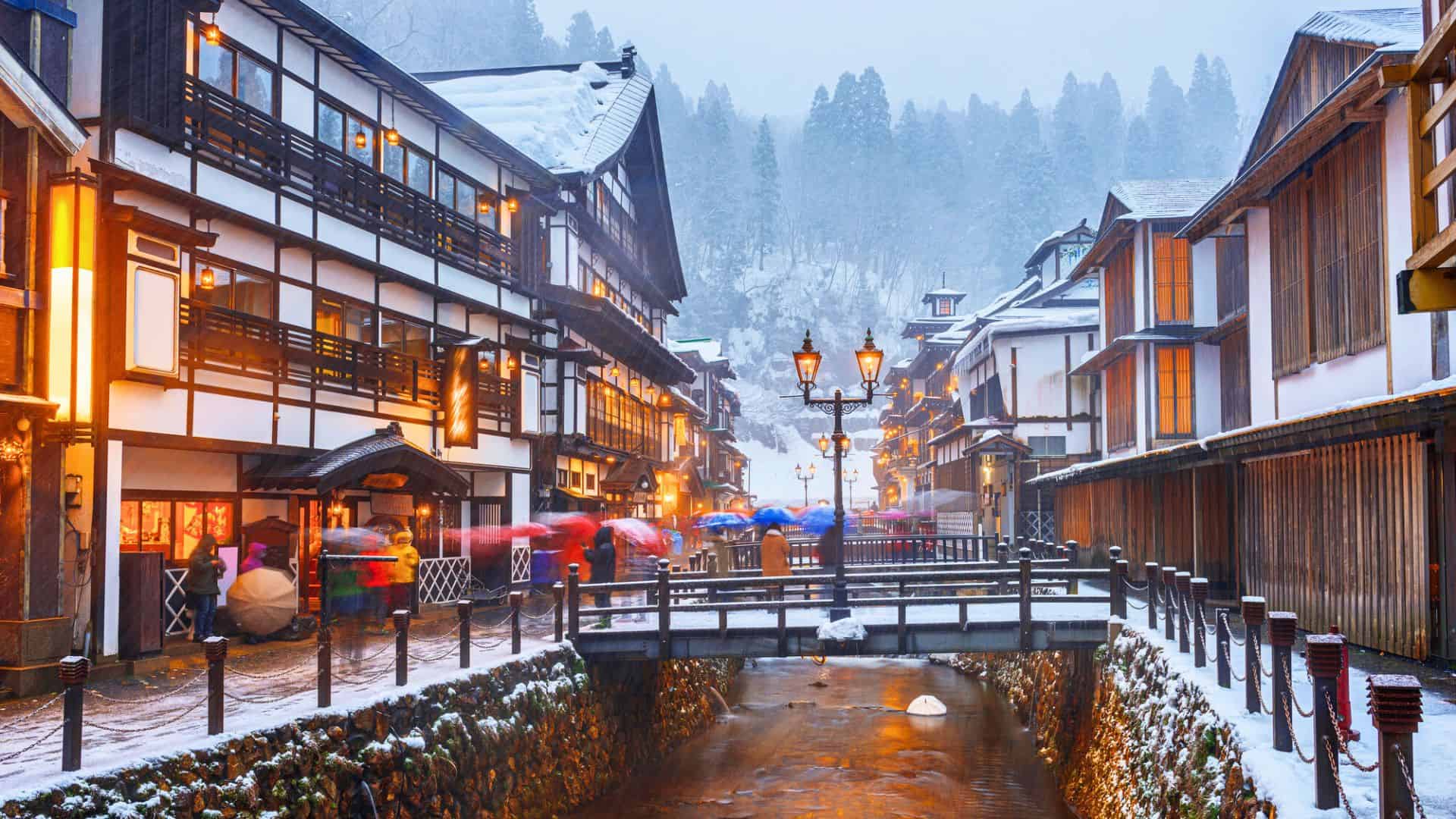 Snow-covered traditional Japanese street with wooden buildings, warm lights, and people with umbrellas on a bridge.