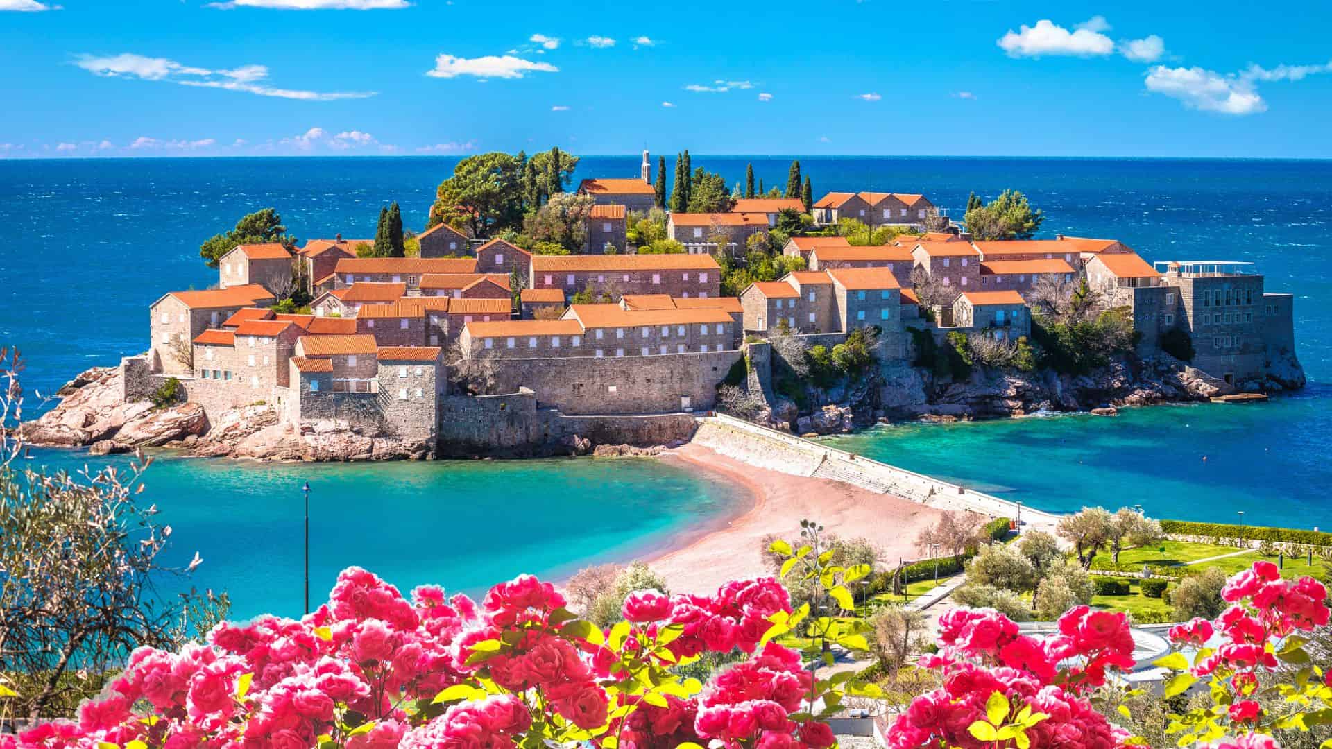 Island village with stone buildings and red roofs, surrounded by blue sea and pink flowers in foreground.