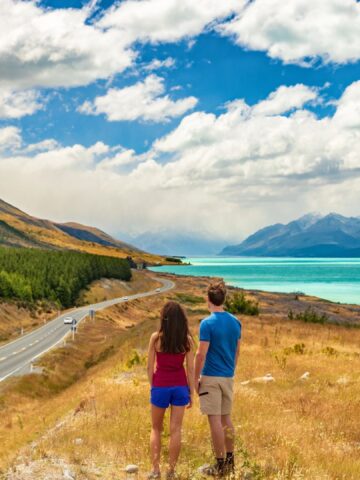 A couple stands by a lakeside road, facing mountains and turquoise water under a partly cloudy sky.