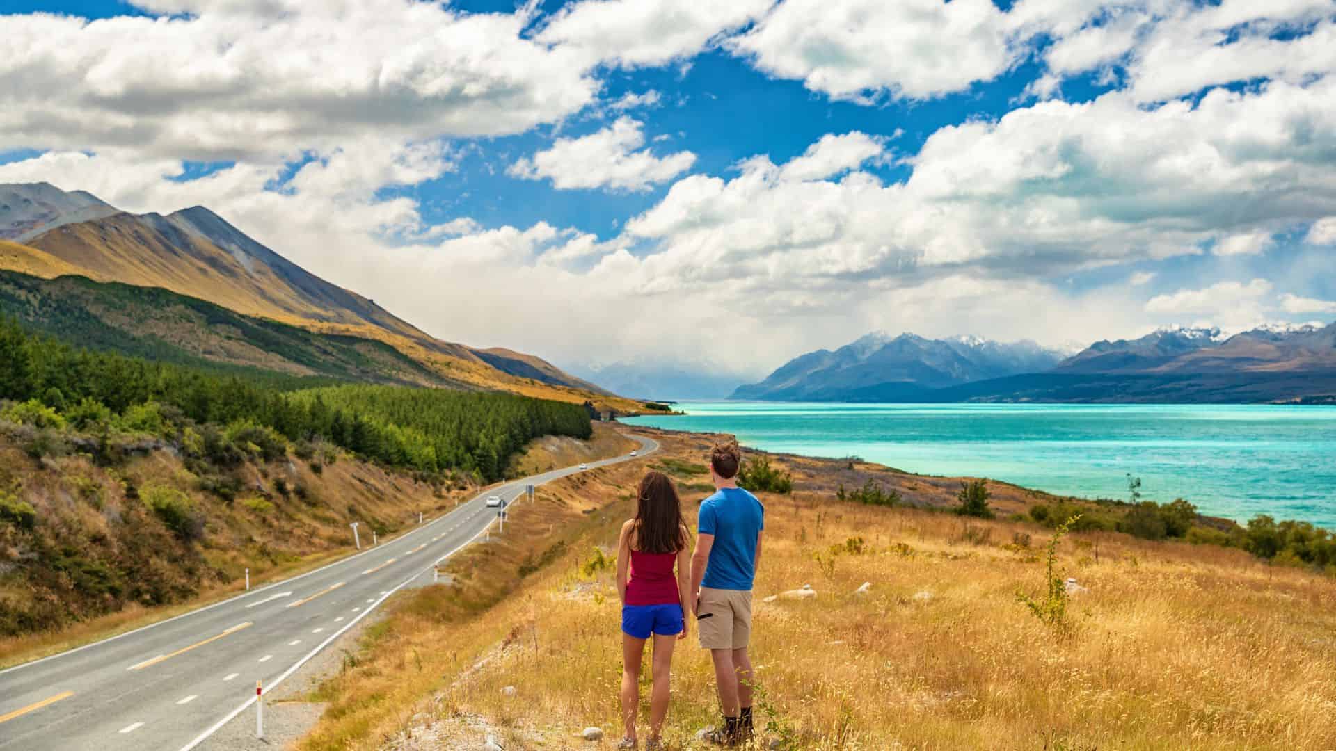 A couple stands by a lakeside road, facing mountains and turquoise water under a partly cloudy sky.