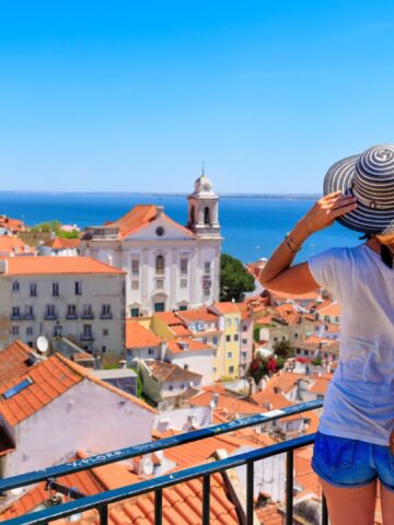 Woman in hat overlooking Lisbon's red rooftops and the sea on a sunny day from a viewpoint.