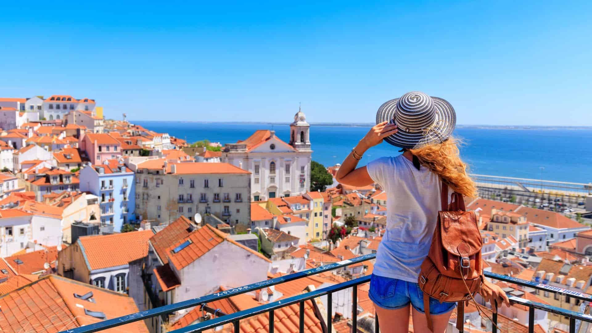 Woman in hat overlooking Lisbon's red rooftops and the sea on a sunny day from a viewpoint.