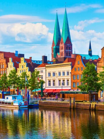 Colorful historic buildings and church spires reflect on a canal in a European city under a blue sky.
