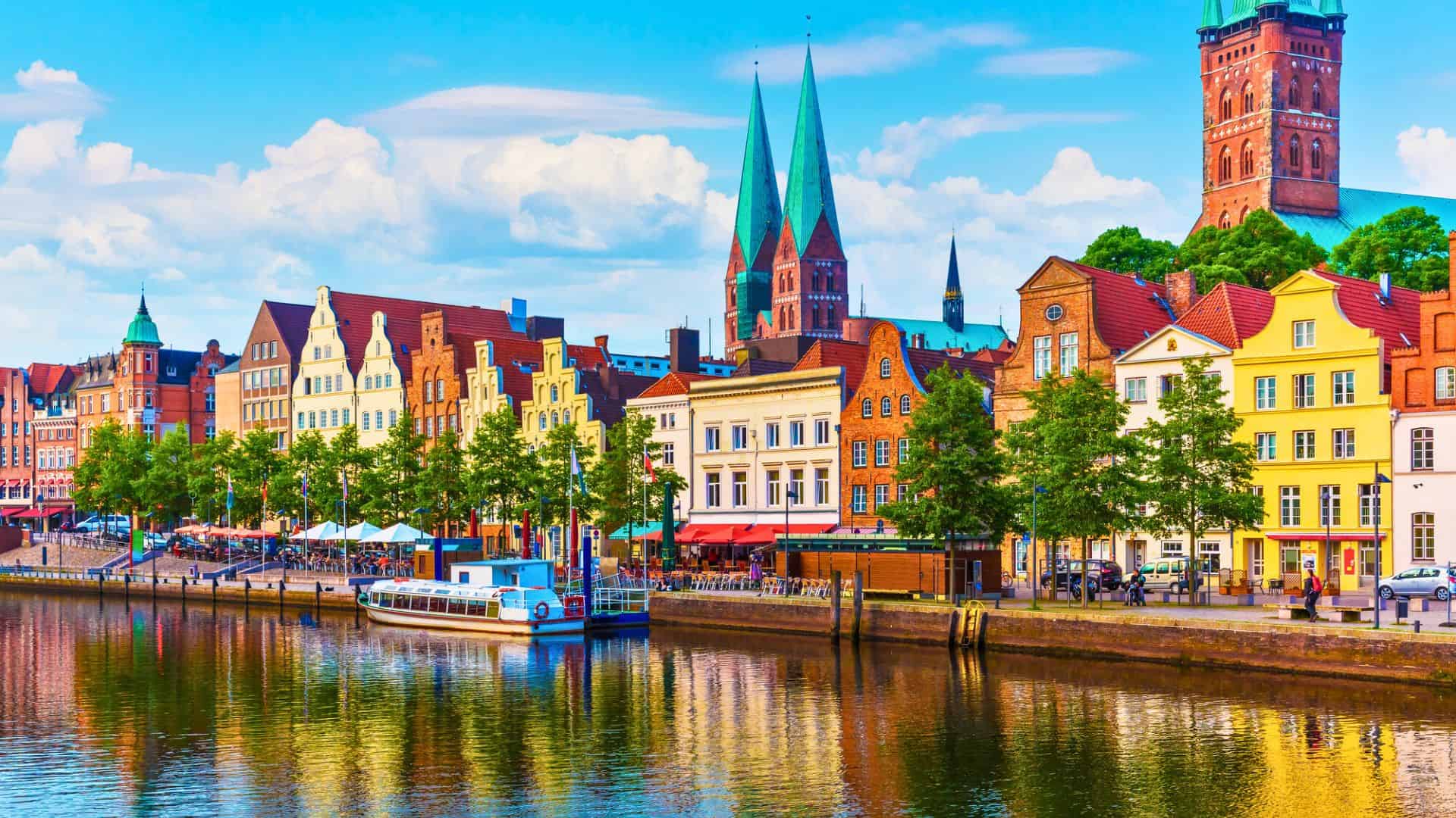 Colorful historic buildings and church spires reflect on a canal in a European city under a blue sky.