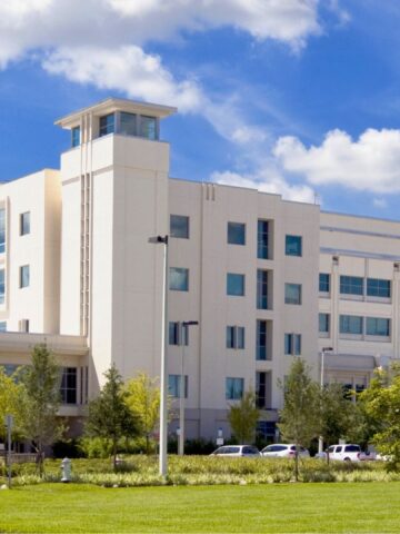 A modern hospital building with large windows, surrounded by trees and grass under a blue sky with clouds.