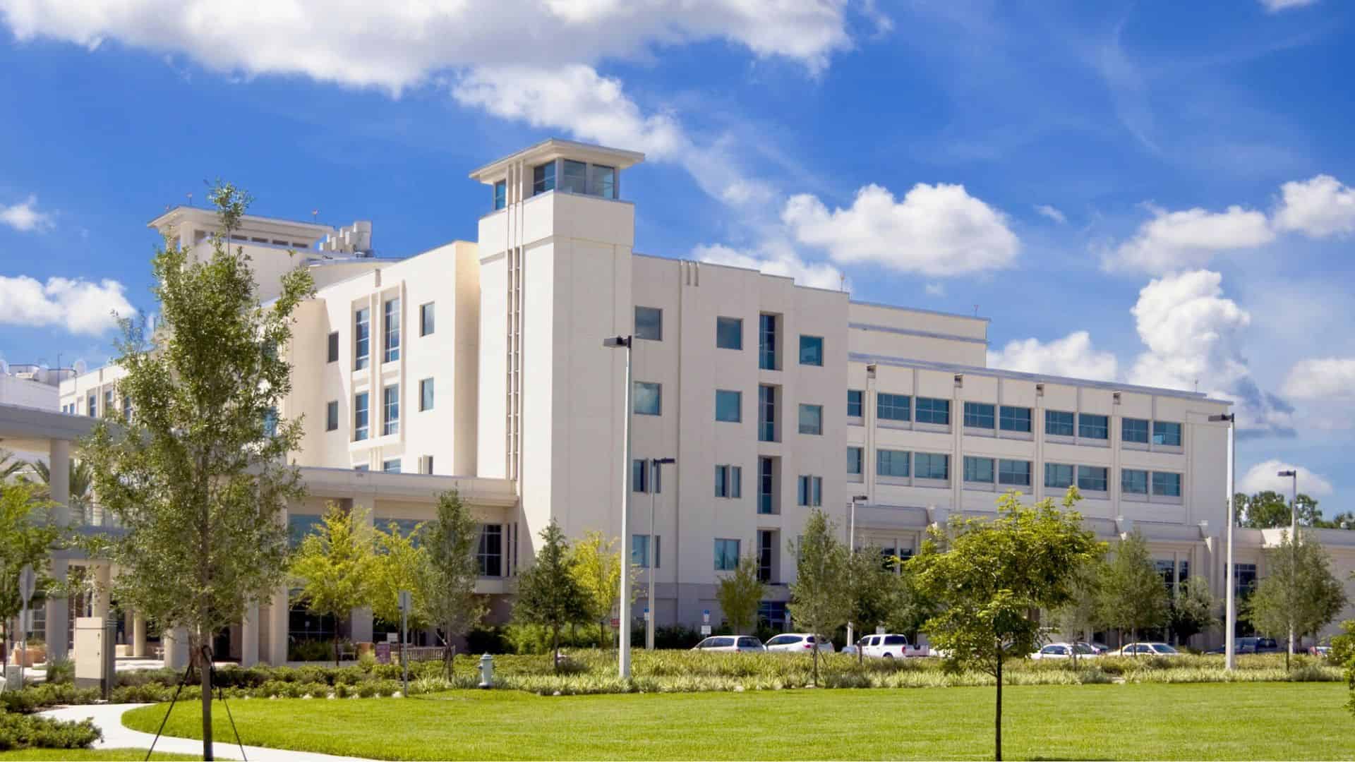 A modern hospital building with large windows, surrounded by trees and grass under a blue sky with clouds.