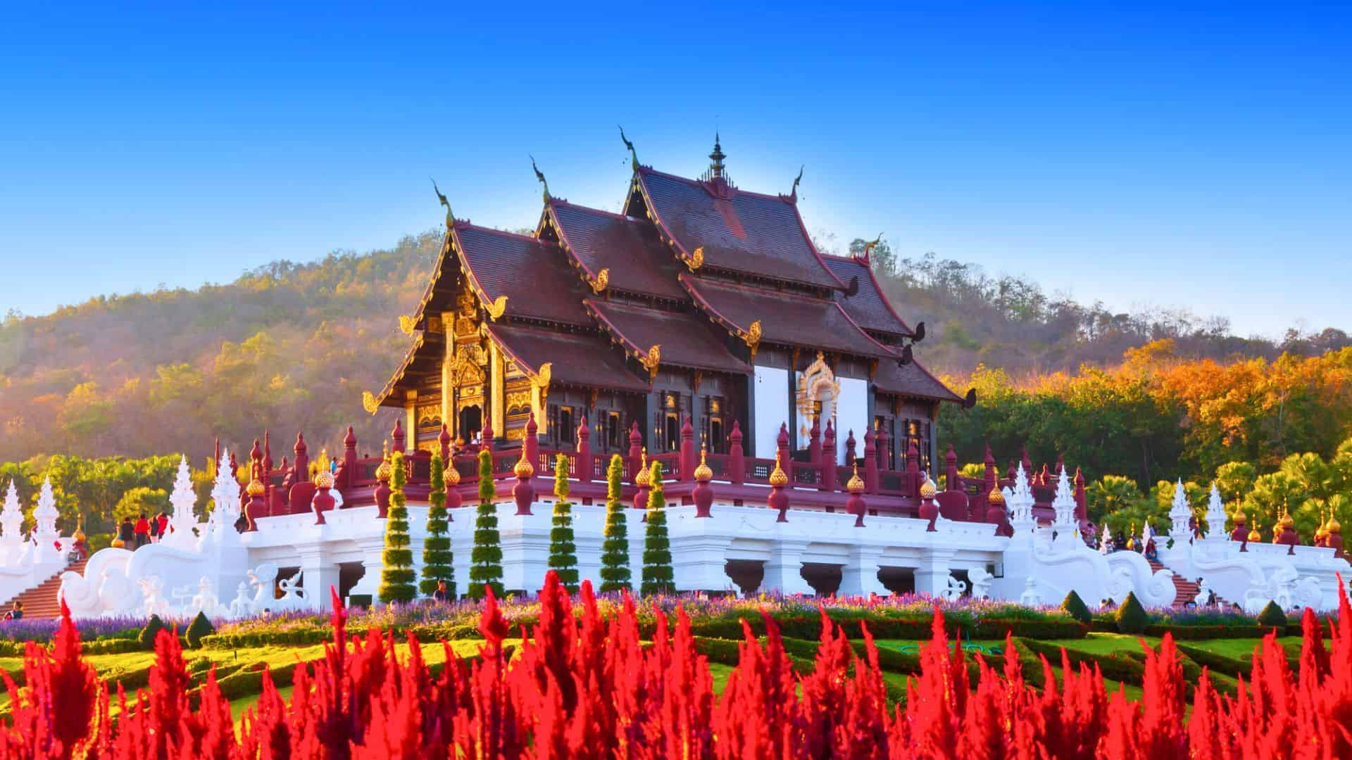 Traditional Thai pavilion with ornate roof, surrounded by red flowers and greenery, set against a blue sky.