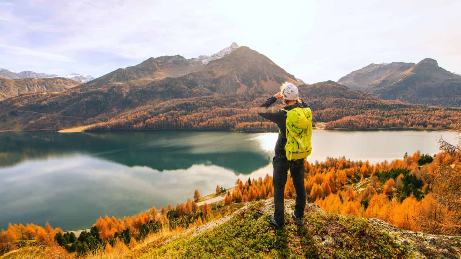 Person with a yellow backpack admires a lake and autumn mountains under a bright sky.